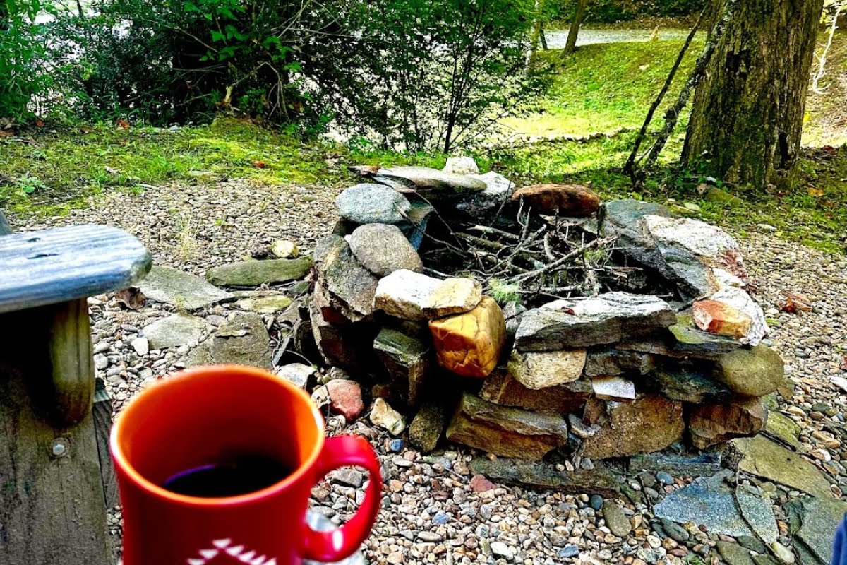 A red mug sits on a wooden surface in focus in the foreground, with an unfocused background showing a rustic stone fire pit surrounded by greenery, highlighting the benefits of nature.