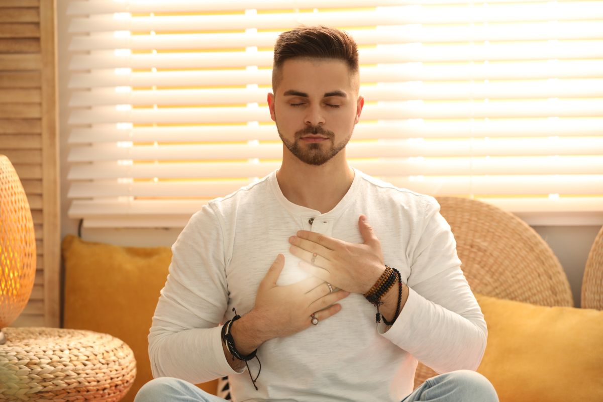 A man meditating with his eyes closed and hands placed on his chest while sitting indoors during a digital detox.