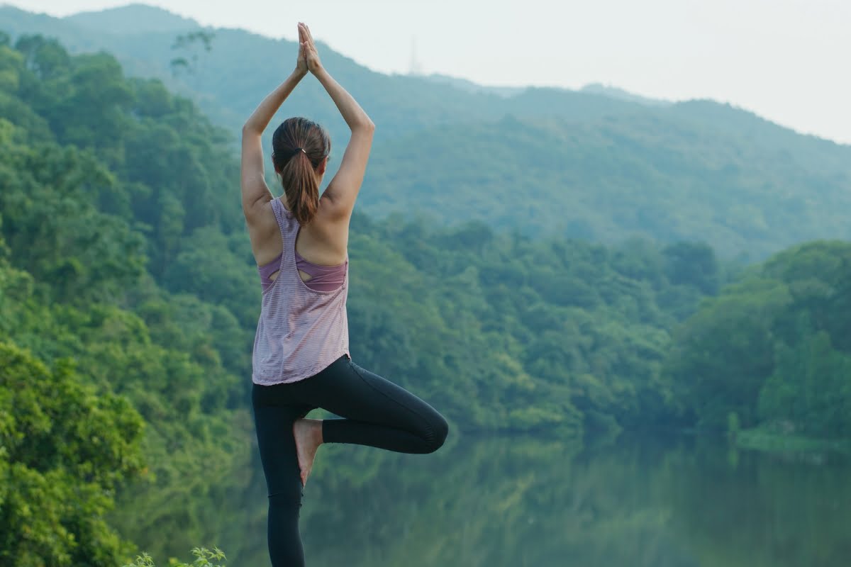 Woman practicing yoga outdoors with arms raised in a tree pose, surrounded by a serene natural landscape, embracing the healing power of Mother Nature.