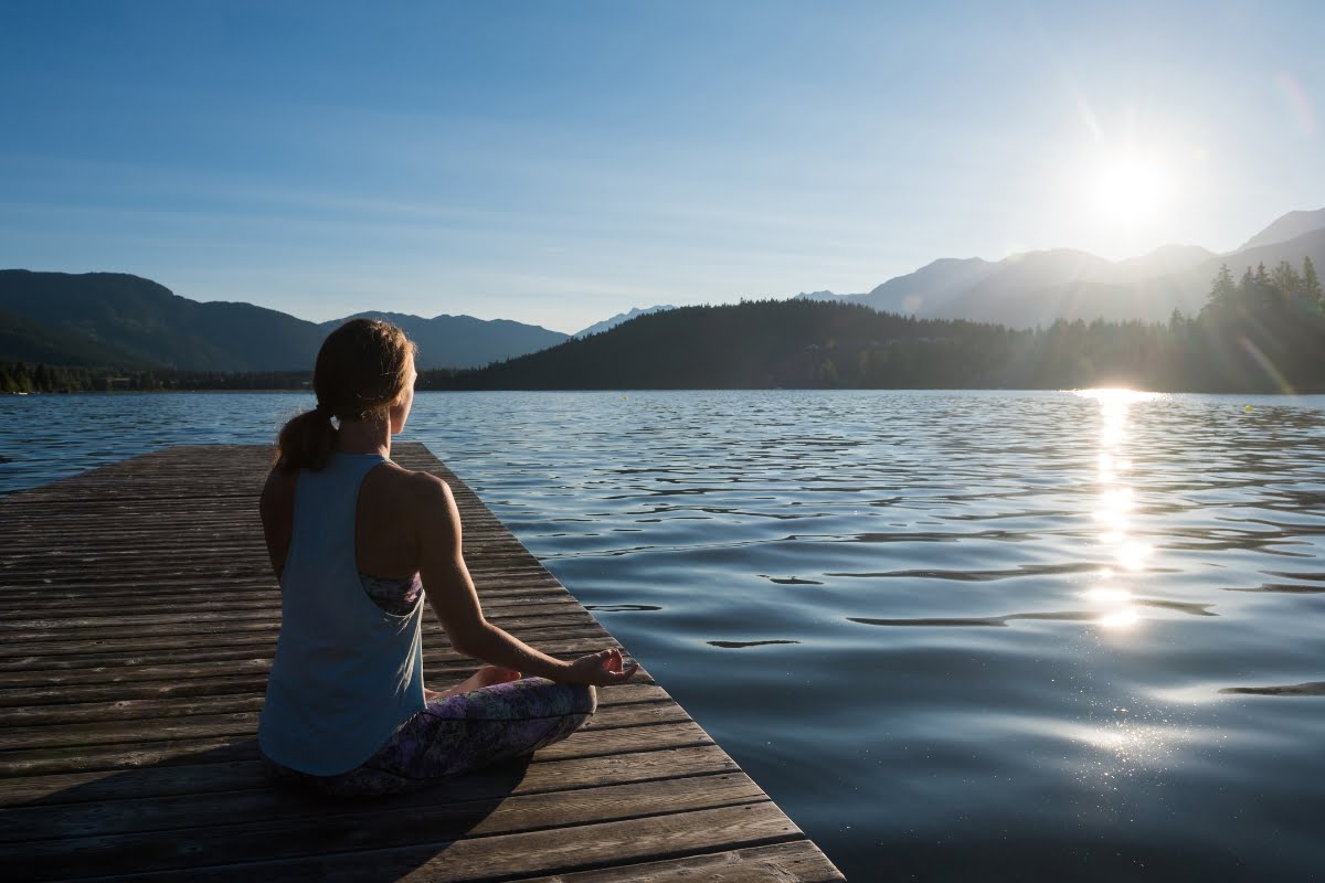 A person sitting on a wooden dock by a lake, enjoying the sunset with mountains in the background experiences the benefits of nature.