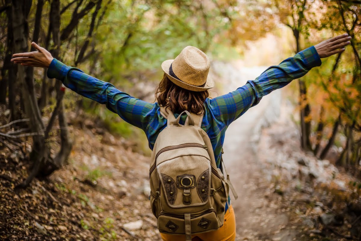 Person with a backpack and hat standing with arms outstretched in a wooded area, embracing the benefits of nature.