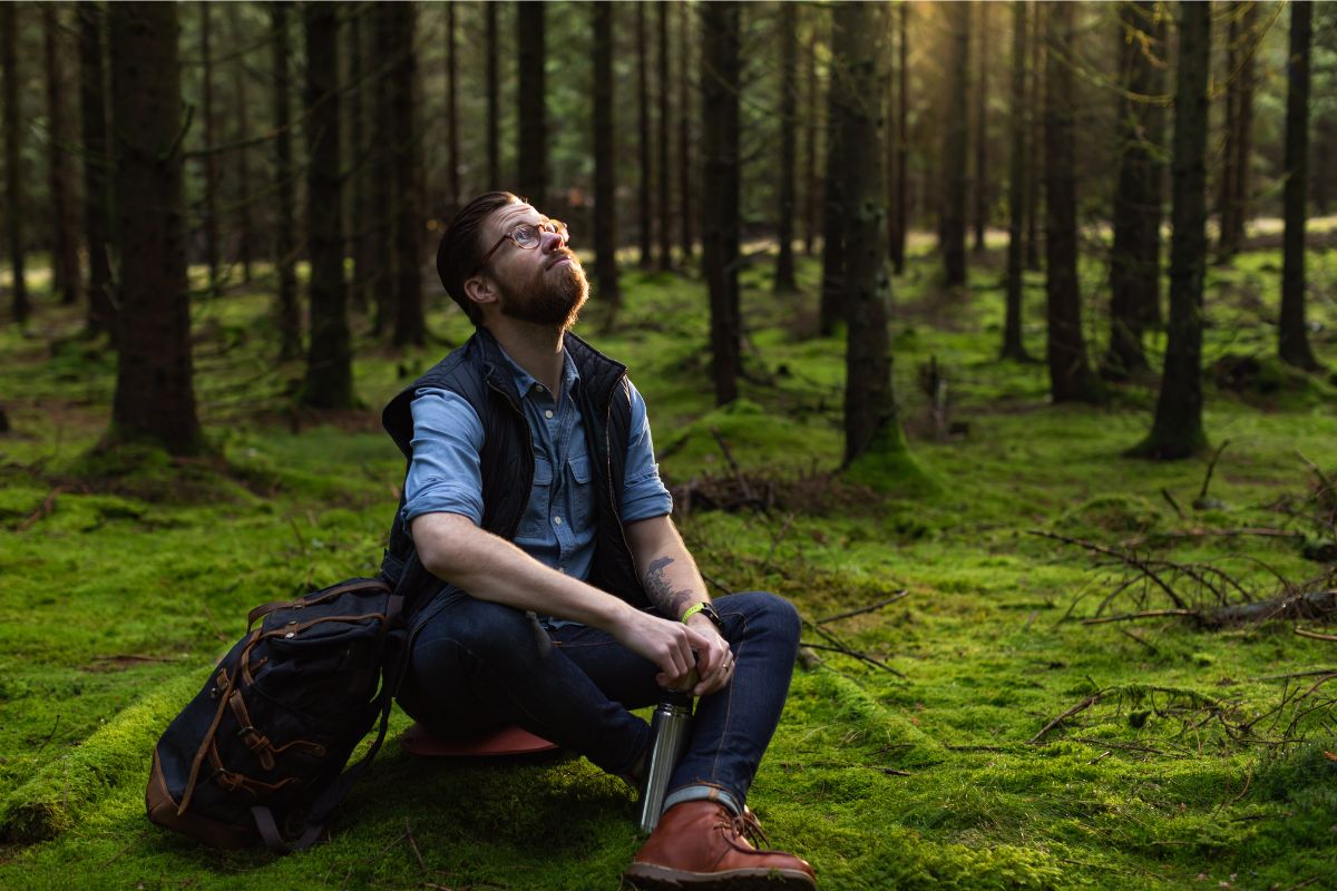 Man practicing relaxation techniques sitting on a red mat in the forest with sunlight filtering through the trees, looking upwards thoughtfully.