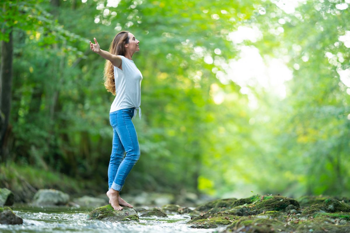 Woman balancing on a rock by a stream with arms outstretched amidst green foliage, practicing nature therapy relaxation techniques at Basecamp Smoky Mountains nature retreats.