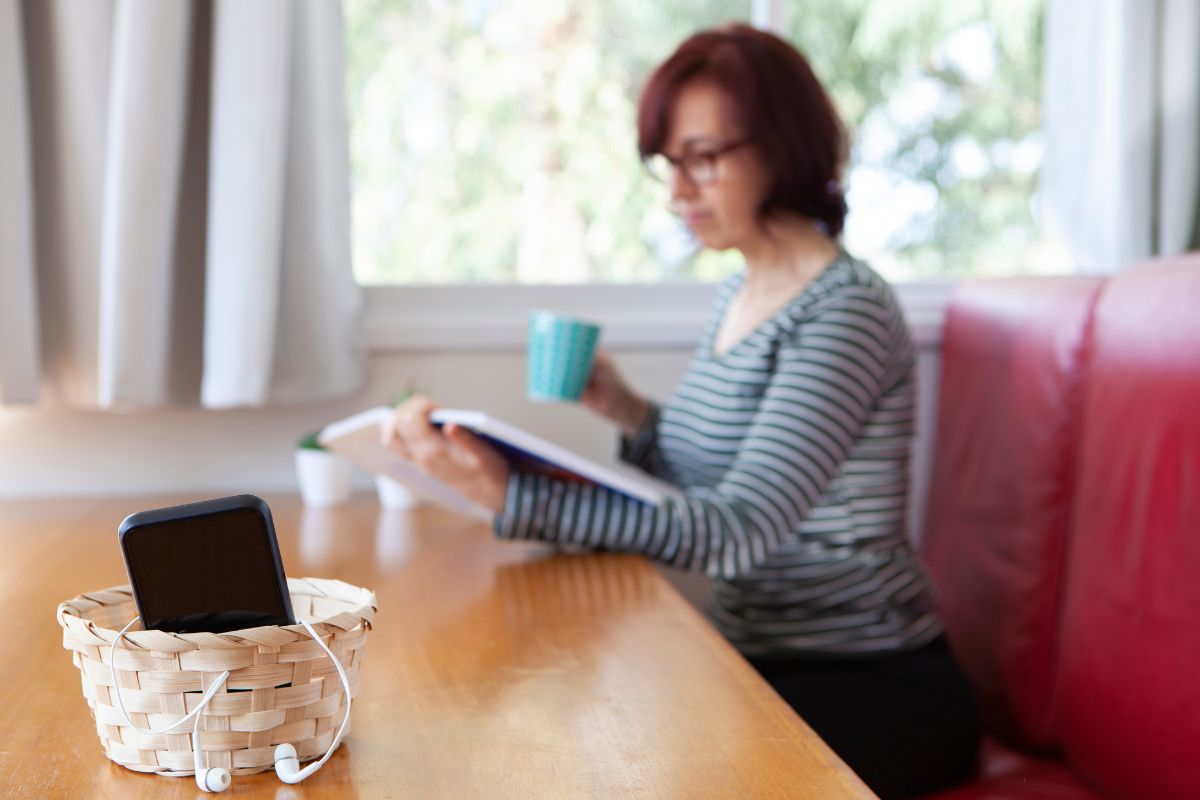 Woman reading a book during a digital detox while holding a mug, with a smartphone in a woven basket on the table in focus.