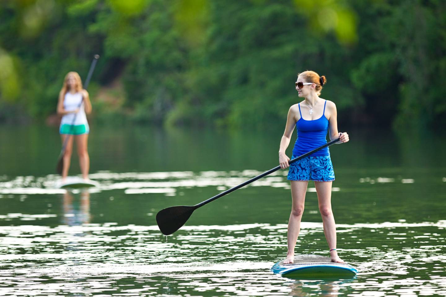 Two women Stand Up Paddleboarding on a calm river, one in the foreground holding a paddle, enjoying the serene experiences with lush greenery in the background.