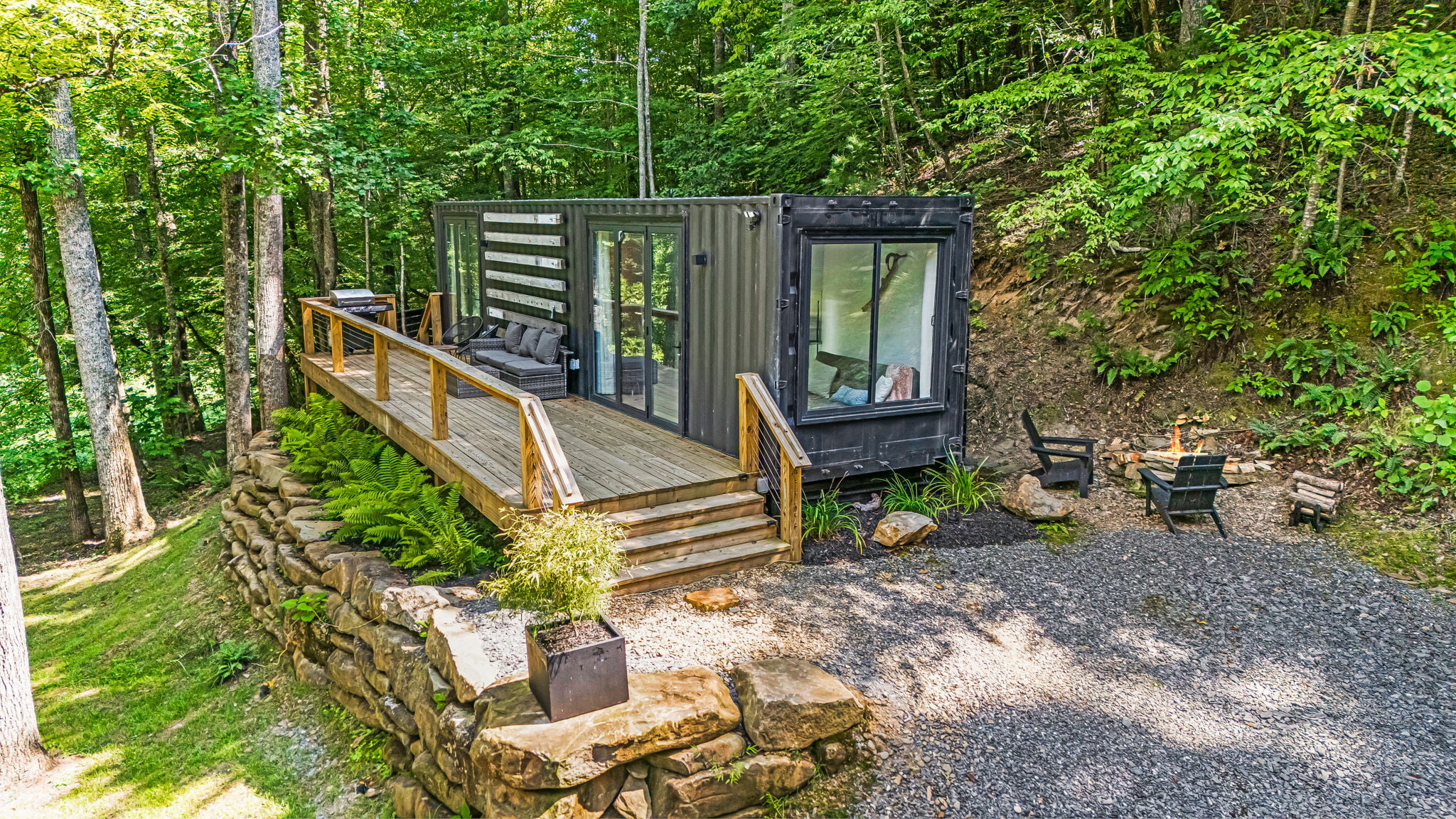 The Elevation Villa, a modern small home built from a shipping container, is situated in the forest. It features a wooden deck with seating and a gravel area with chairs, all amidst lush greenery for an idyllic retreat.