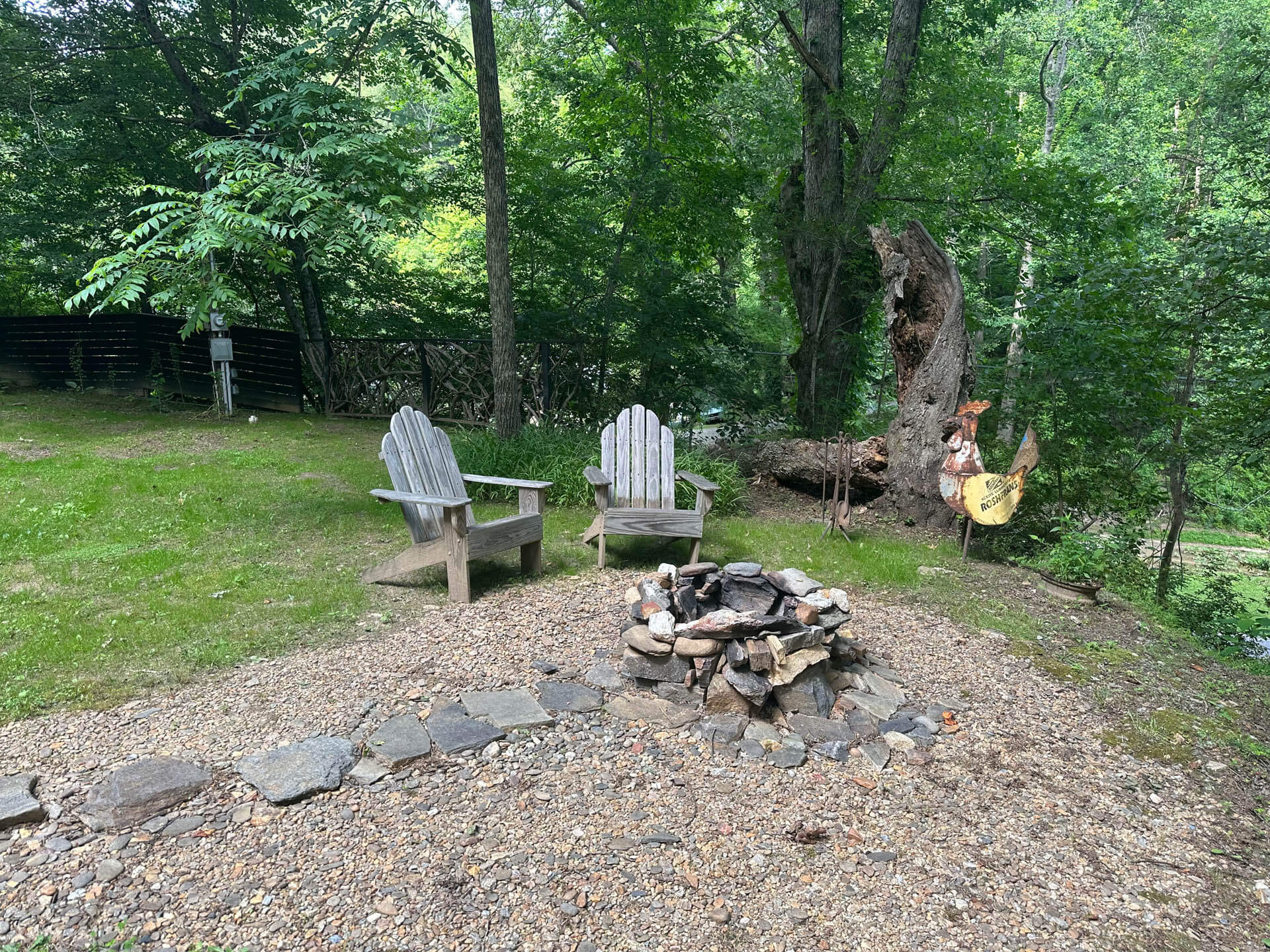 Two wooden adirondack chairs facing a stone fire pit, with a large hollow tree and lush greenery in the background.