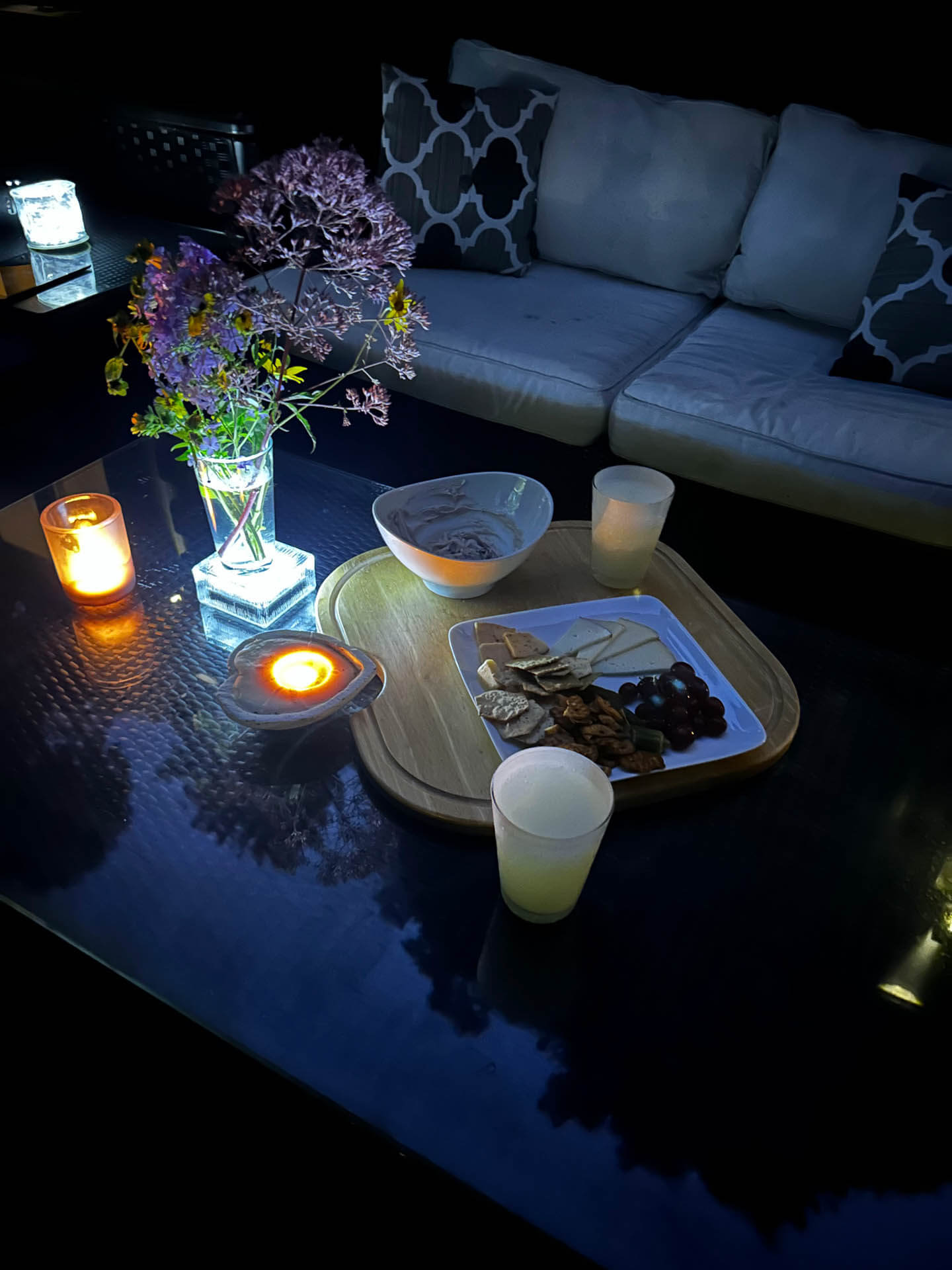 A cozy living room scene at night with a sofa and coffee table, adorned with candles, snacks, and a vase of flowers, illuminated by ambient blue lighting.