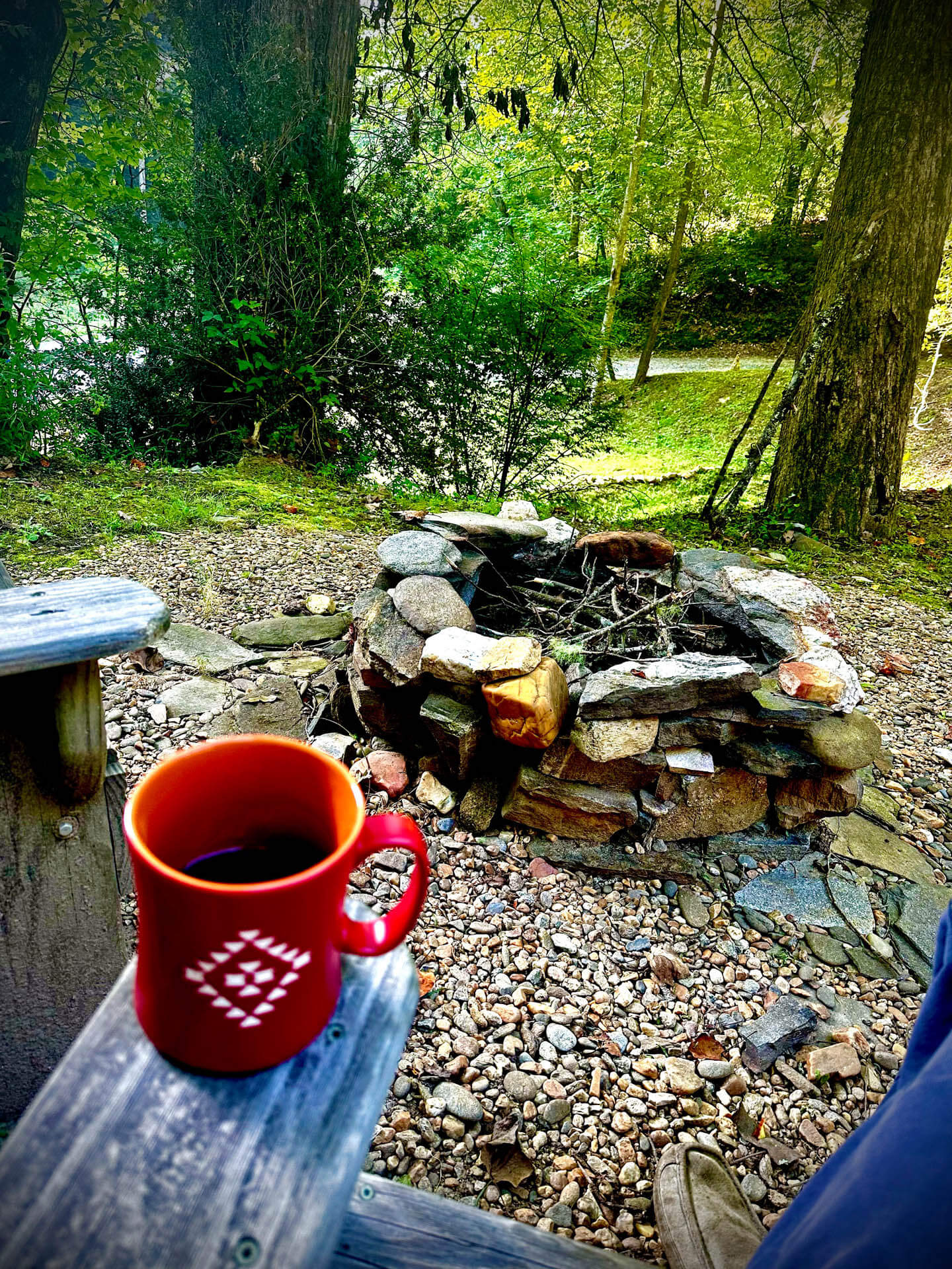 A red mug sits on a wooden railing overlooking an unlit stone fire pit surrounded by trees and gravel.