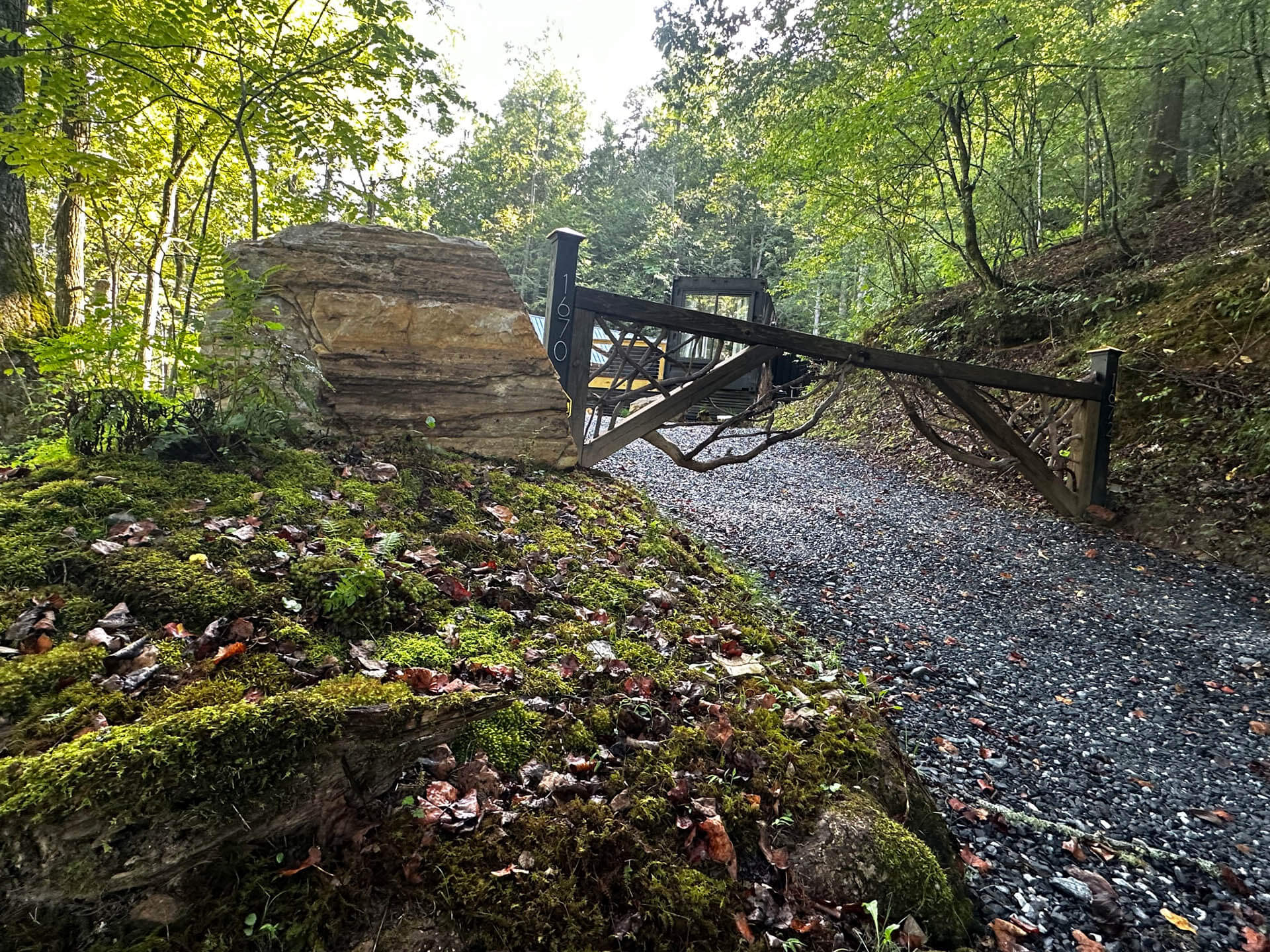 A rustic wooden barrier blocks a gravel path in a lush, green forest, with sunlight filtering through the trees.