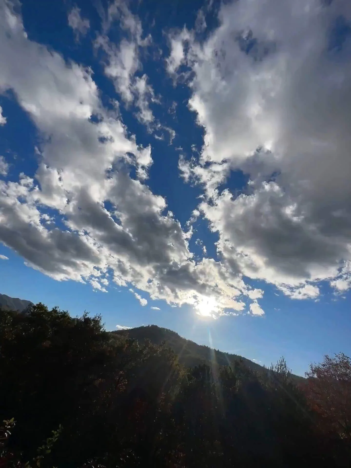 Sun shining through scattered clouds over a mountainous landscape with rays casting light on the foliage below.