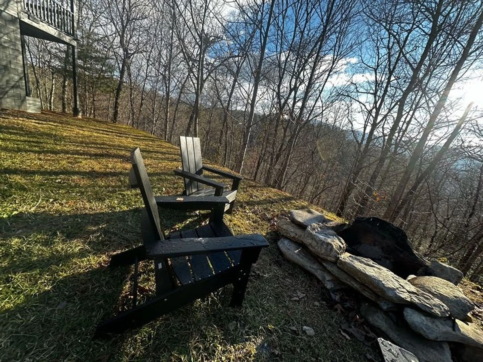 Two wooden chairs on a grassy hillside facing a view of bare trees and distant hills, with a low stone wall and a balcony in the background.