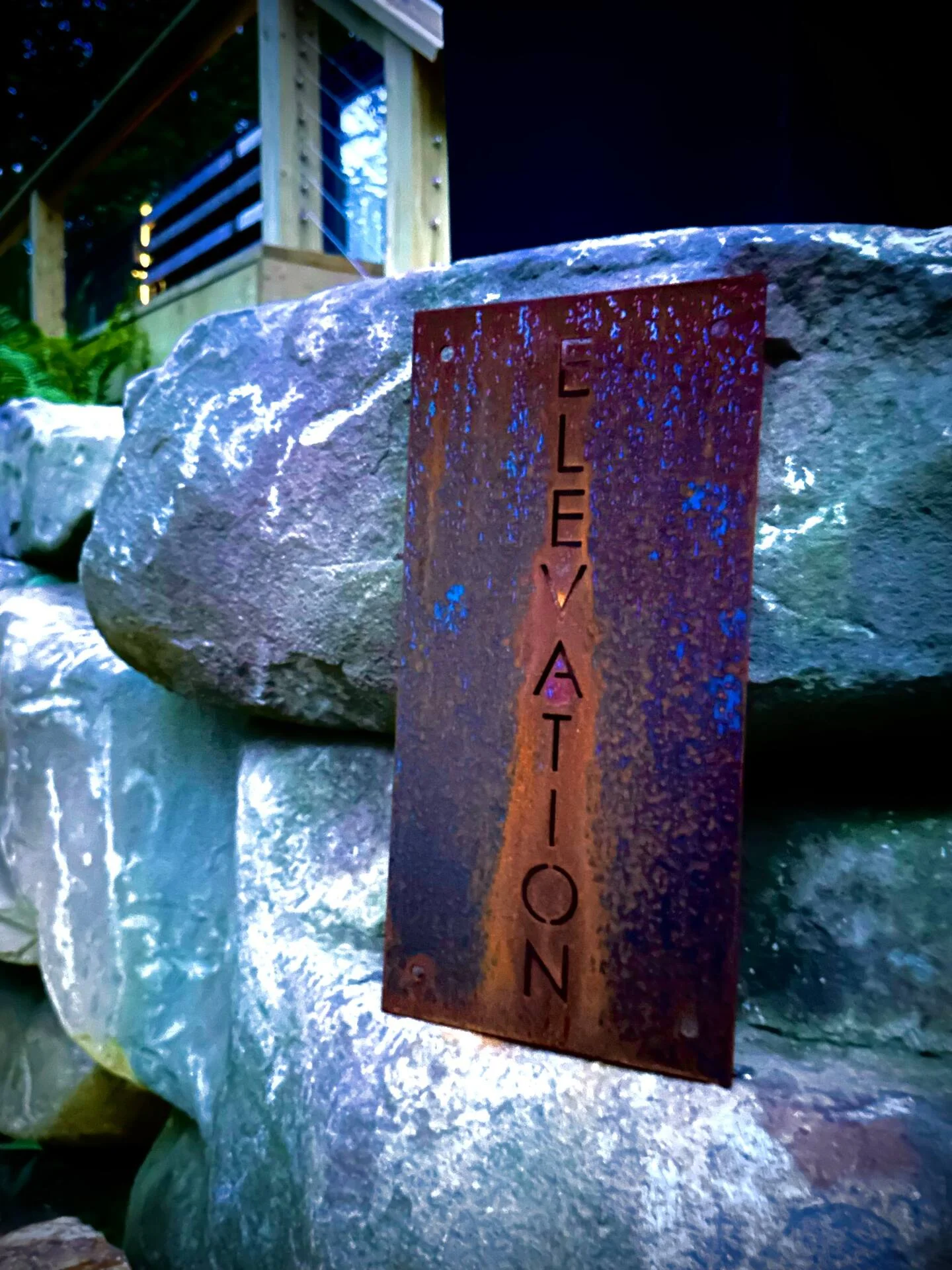 Rusted metal sign labeled "elevation" mounted on a stack of blue-lit boulders at dusk.