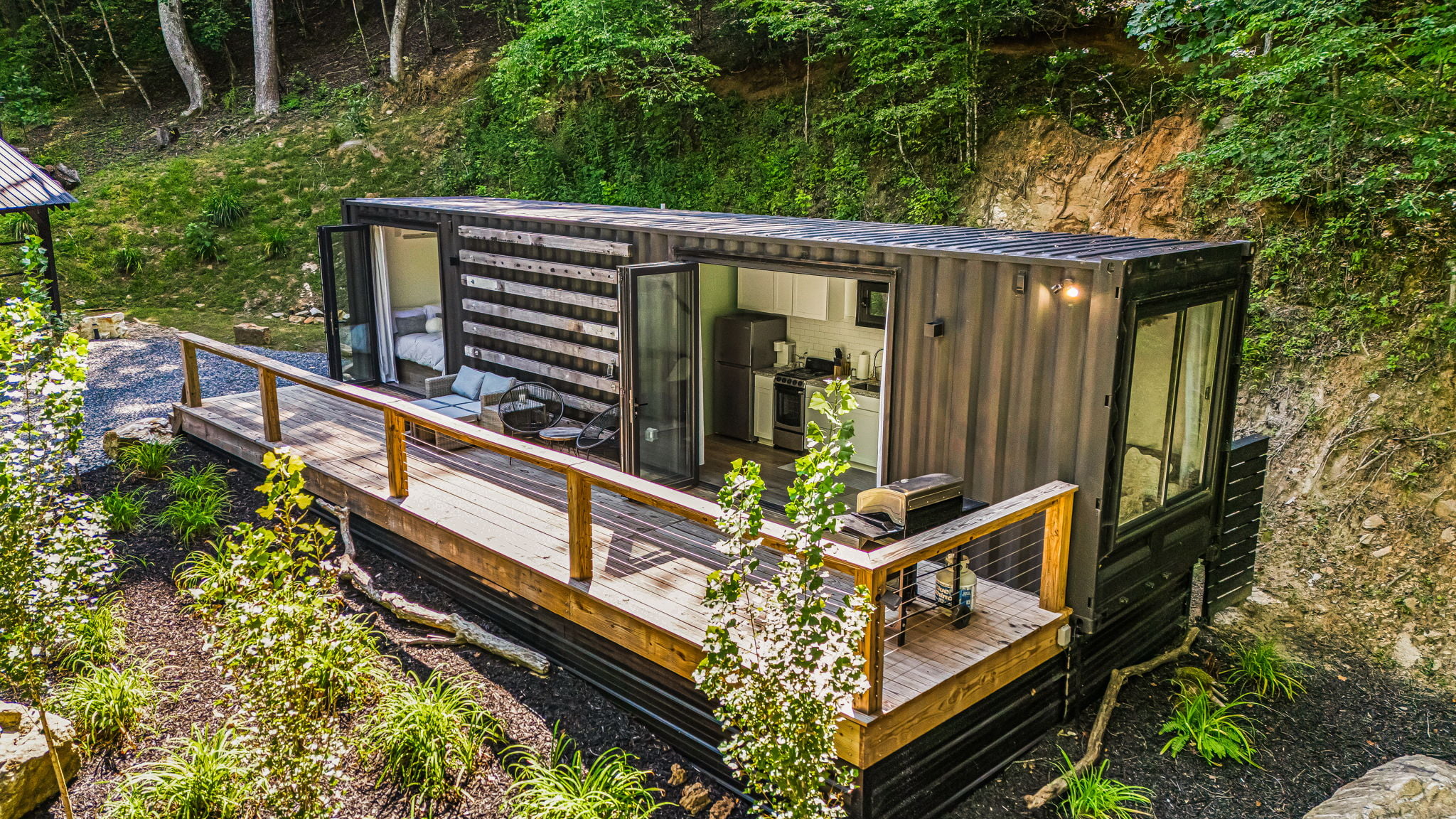 A modern container home with large glass doors, a wooden deck, outdoor seating, and surrounded by greenery. Welcome to Tabs 02, a grounded villa that seamlessly blends nature and contemporary design.