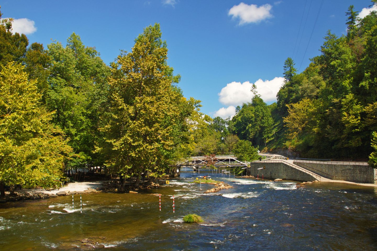 A scenic view of a river with fast-moving water, surrounded by lush green trees under a clear blue sky, featuring local attractions including a pedestrian bridge in the background.