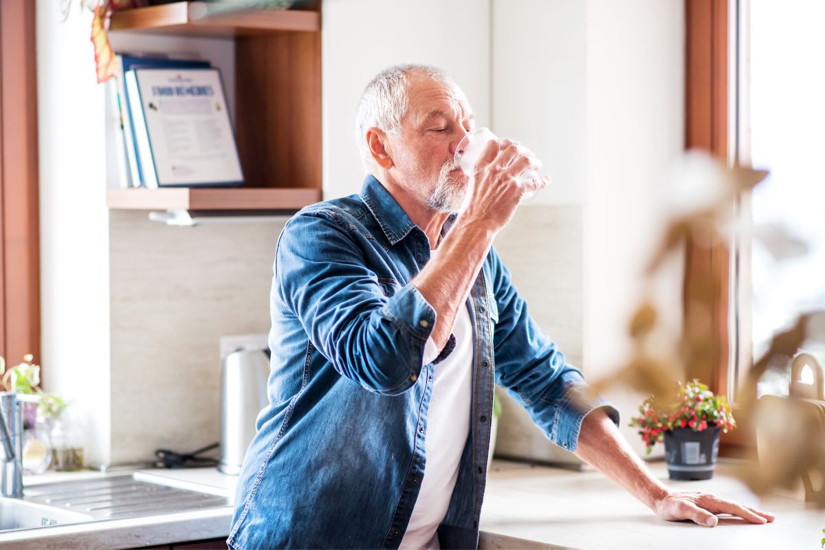 Senior man in a denim shirt experiencing the healing power of water, drinking from a glass in a sunny kitchen.