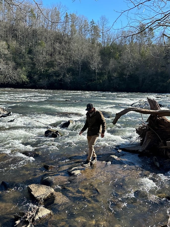 A person wades across a rocky stream with rapid flowing water, surrounded by trees under a clear blue sky. The provided text "About" is too brief and lacks context or specific content to extract or determine important SEO keywords. Please provide a more detailed description for accurate keyword extraction.