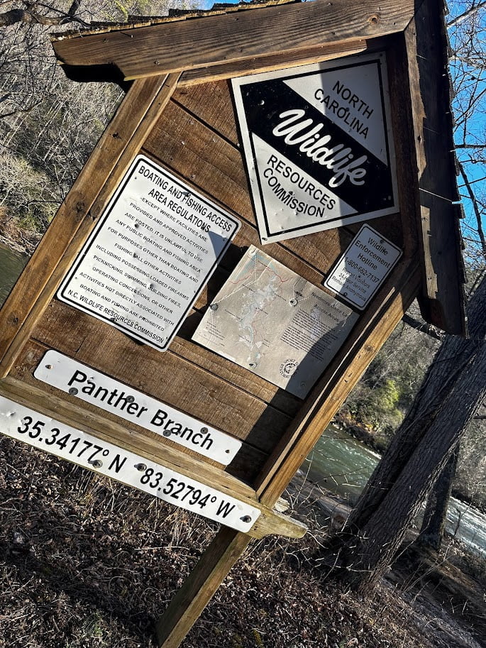 Wooden signboard titled "Panther Branch" with maps and regulatory information, by a riverbank, under clear skies.