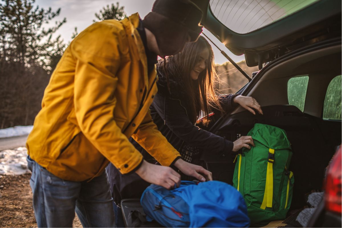 Two people packing a green and yellow backpack into the trunk of a car during a sunset woodland adventure travel.