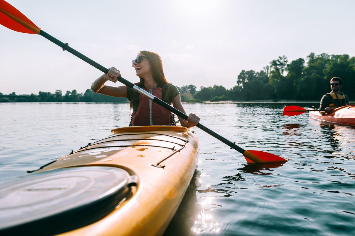 A woman kayaking on a serene lake, smiling, with a man in another kayak in the background, enjoying the latest adventure travel trend on a bright sunny day.