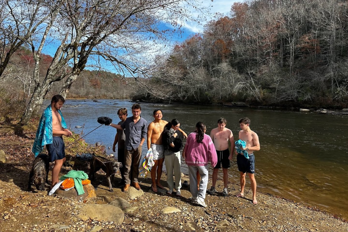 A group of young people in swimwear stand by a riverbank with trees, some with autumn foliage, under a clear sky, enjoying the day's experiences.