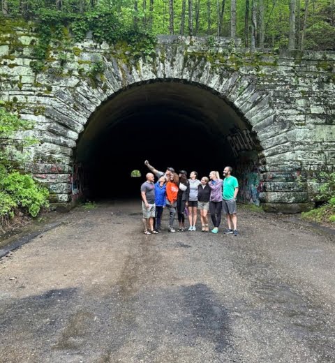 Group of seven people posing for a photo in front of a large, arched stone tunnel entrance surrounded by trees, ready for their hiking adventures.