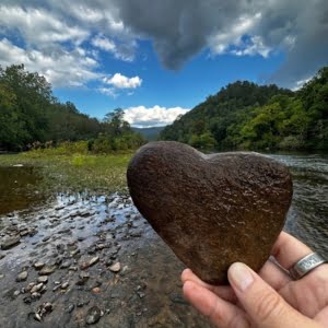 A hand holding a heart-shaped rock with a river and forested hills under a cloudy sky in the background, capturing the essence of hiking adventures.