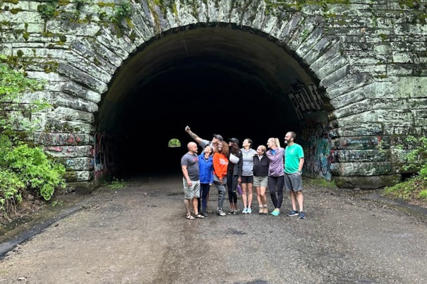 A group of seven friends capturing their vibrant life experiences by posing and smiling in front of a graffiti-covered tunnel entrance, with one person holding a selfie stick.