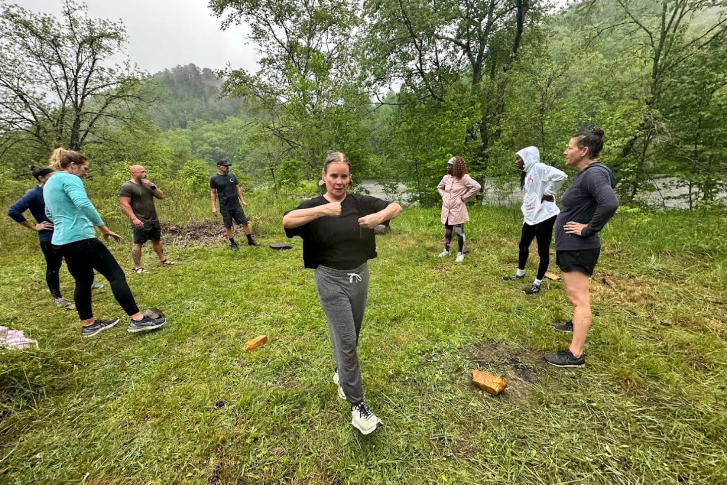 A fitness instructor demonstrates a stretching exercise to a group of adults during an outdoor wellness experience in a rainy, grassy field surrounded by trees.