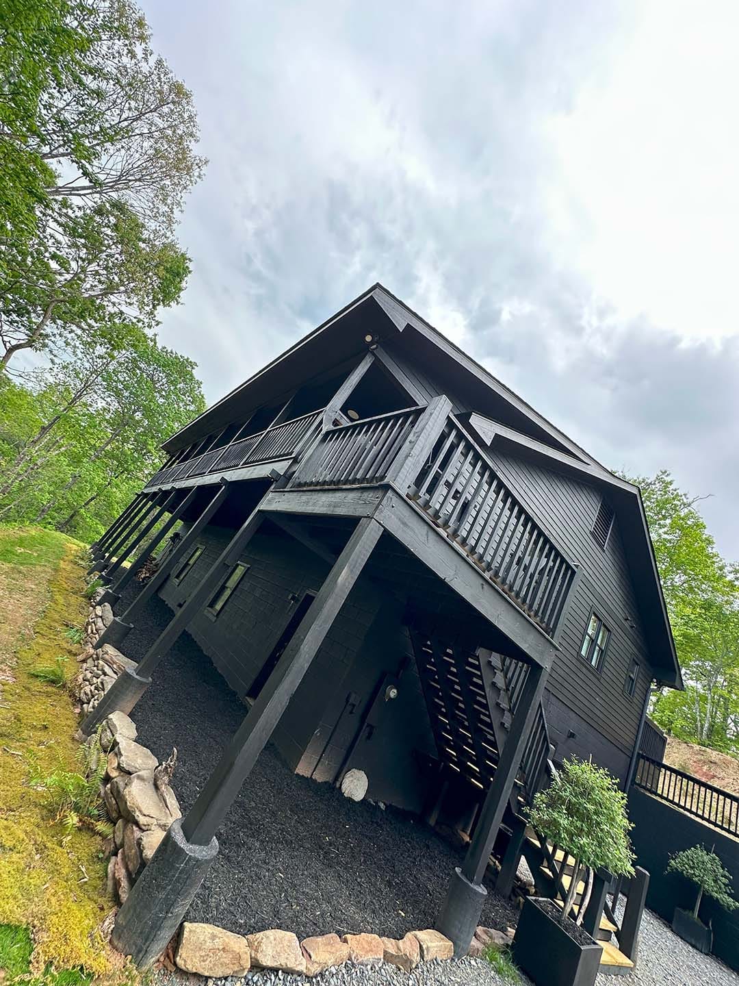 A dark wooden Summit Lodge with a raised deck and stone-bordered landscaping sits on a sloped yard, surrounded by green trees under a cloudy sky.