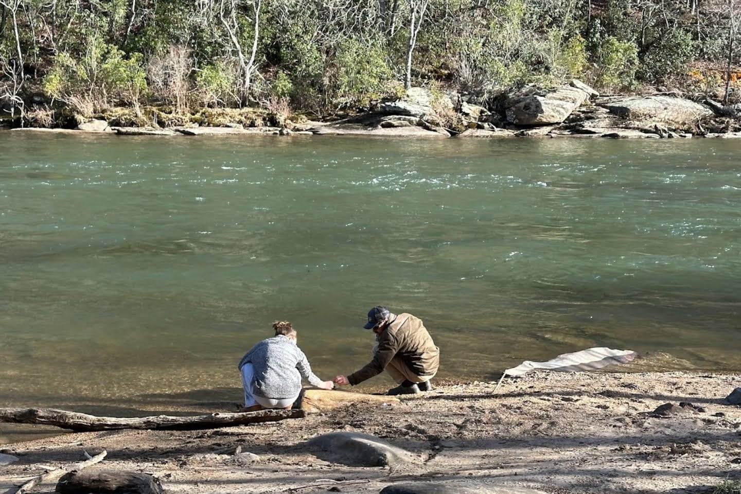 Two people crouch near the edge of a river, immersed in their surroundings. The trees and rocky terrain create a backdrop for their shared experience with nature.