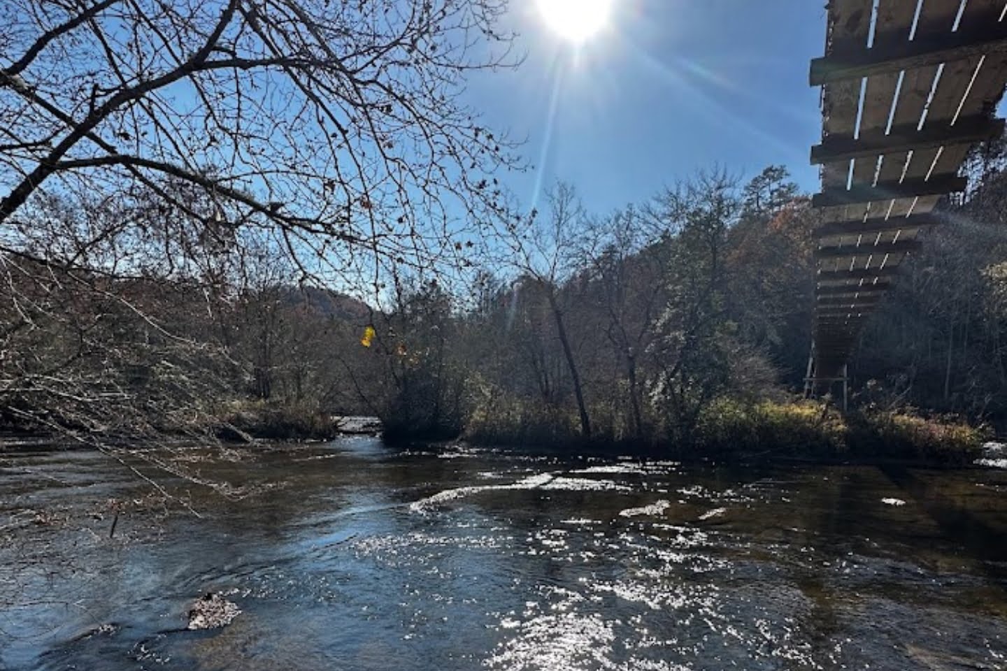 Sunlit river with shimmering water, surrounded by lush trees, under a clear blue sky, featuring a rustic wooden bridge structure on the right.