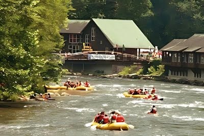 Groups of people experiencing whitewater kayaking on a river in front of a large green-roofed building with a wooden deck, surrounded by trees.