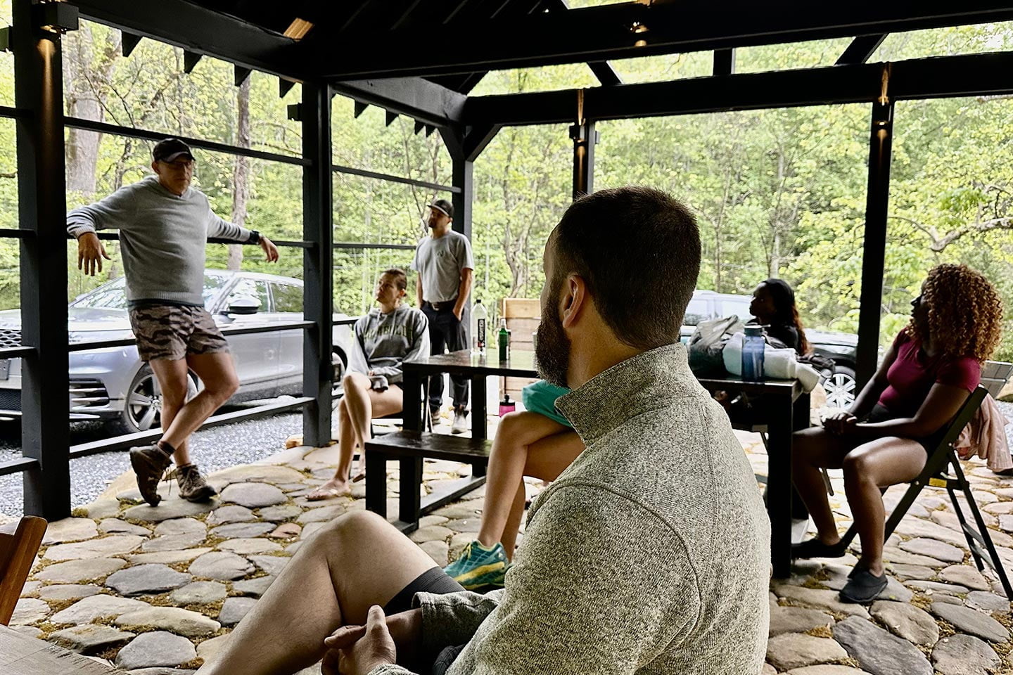 A man gestures while speaking to a group of people seated on a covered patio with a stone floor surrounded by trees, creating an engaging outdoor experience.