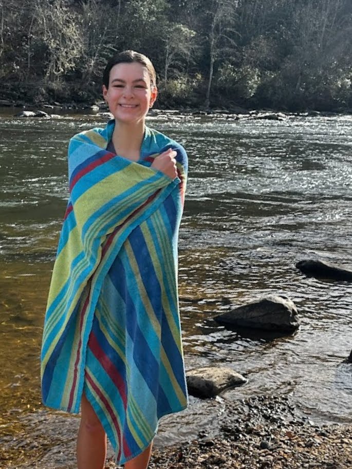 Woman wrapped in a striped towel smiling by a cold plunge pool with sunlight filtering through trees in the background.