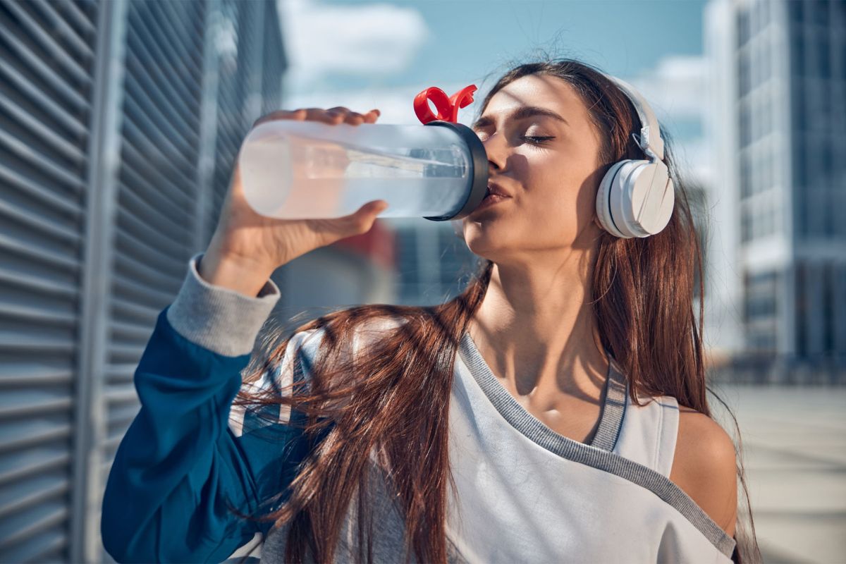 A young woman wearing headphones drinks water from a bottle outdoors, experiencing the healing power of water, with urban buildings in the background.