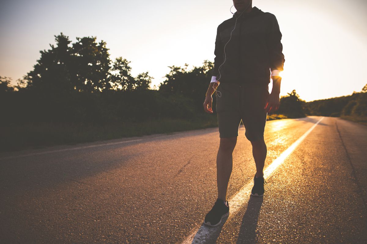 A person practicing the art of mindful walking down a sunlit road at sunset, holding a bottle, dressed in shorts and a jacket.