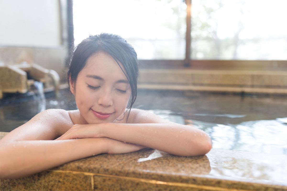 A young woman relaxes with closed eyes in an onsen, resting her arms and chin on the stone edge, experiencing the healing power of water, with a window in the background.