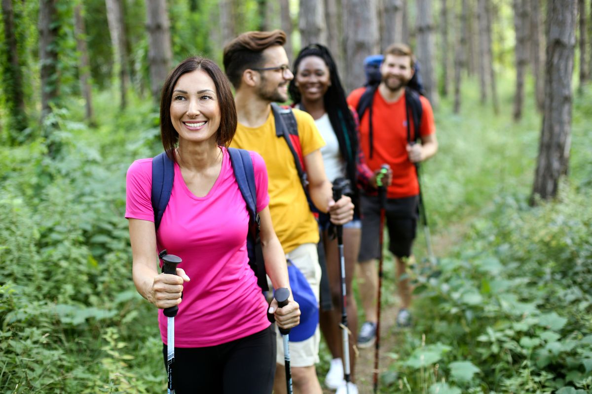 Group of four diverse friends hiking together in a green forest, equipped with backpacks and hiking poles, smiling and enjoying the spring flora.