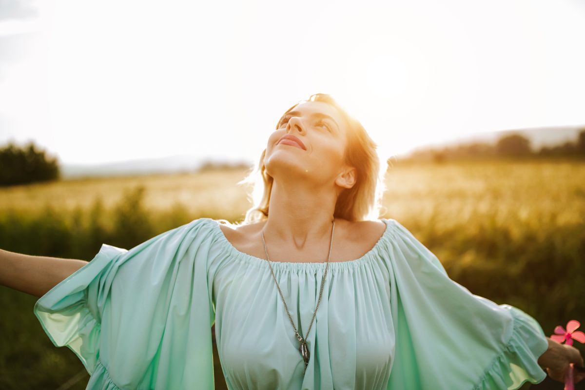 Woman in a flowing green dress standing in a field surrounded by spring flora at sunset, arms outstretched, looking upwards with a peaceful expression.