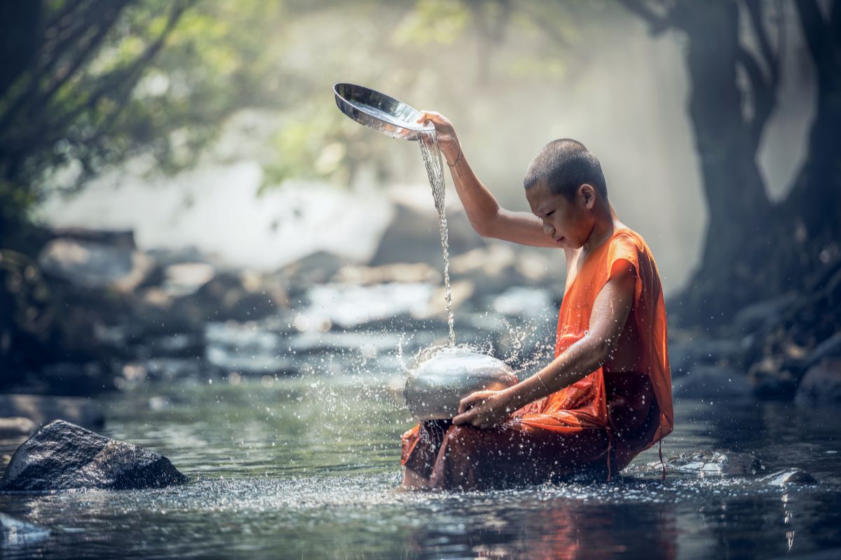 A young monk in orange robes pours water from a bowl to demonstrate the healing power of water, while seated on a rock in a misty, sunlit stream.