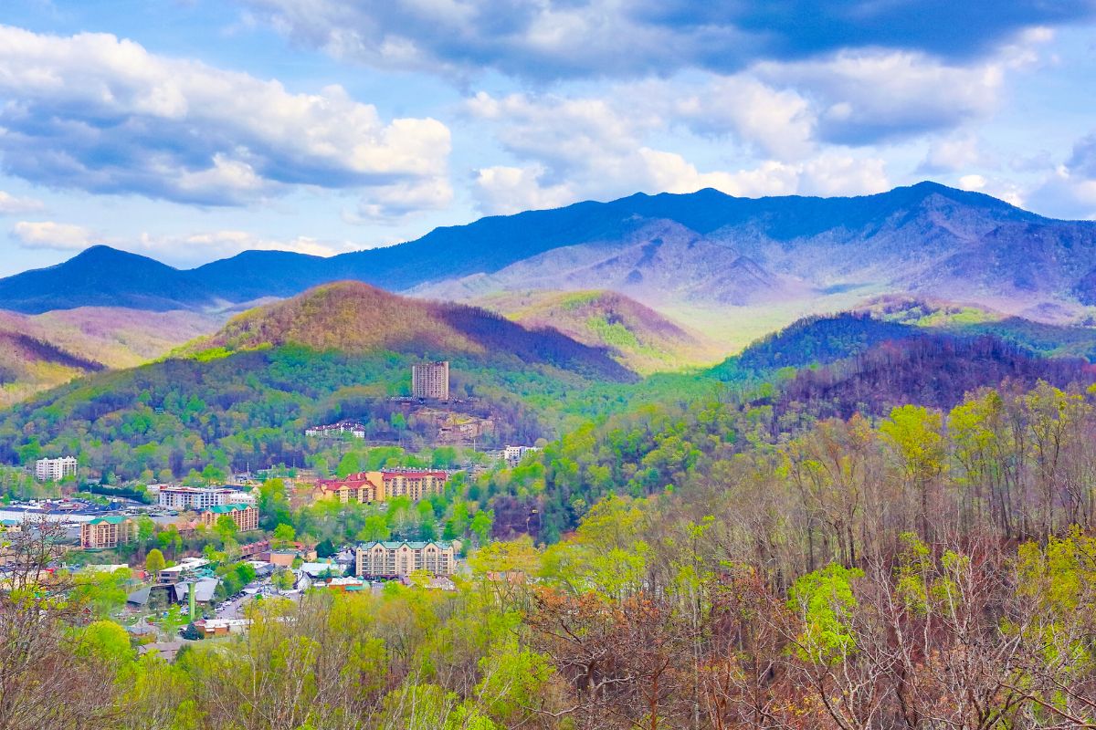 Vibrant aerial view of a town nestled in a valley, surrounded by lush spring flora and fauna and green mountains in the background under a partly cloudy sky.