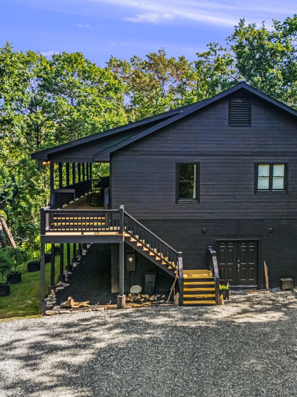 A two-story dark-colored house, reminiscent of the charming Summit Lodge, features an external staircase, a wooden deck, and an elevated porch set amidst lush greenery and gravels.