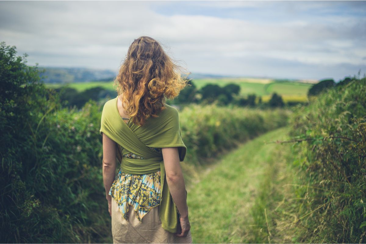 Woman with curly hair practicing the art of mindful walking on a narrow path through lush green fields, viewed from behind.