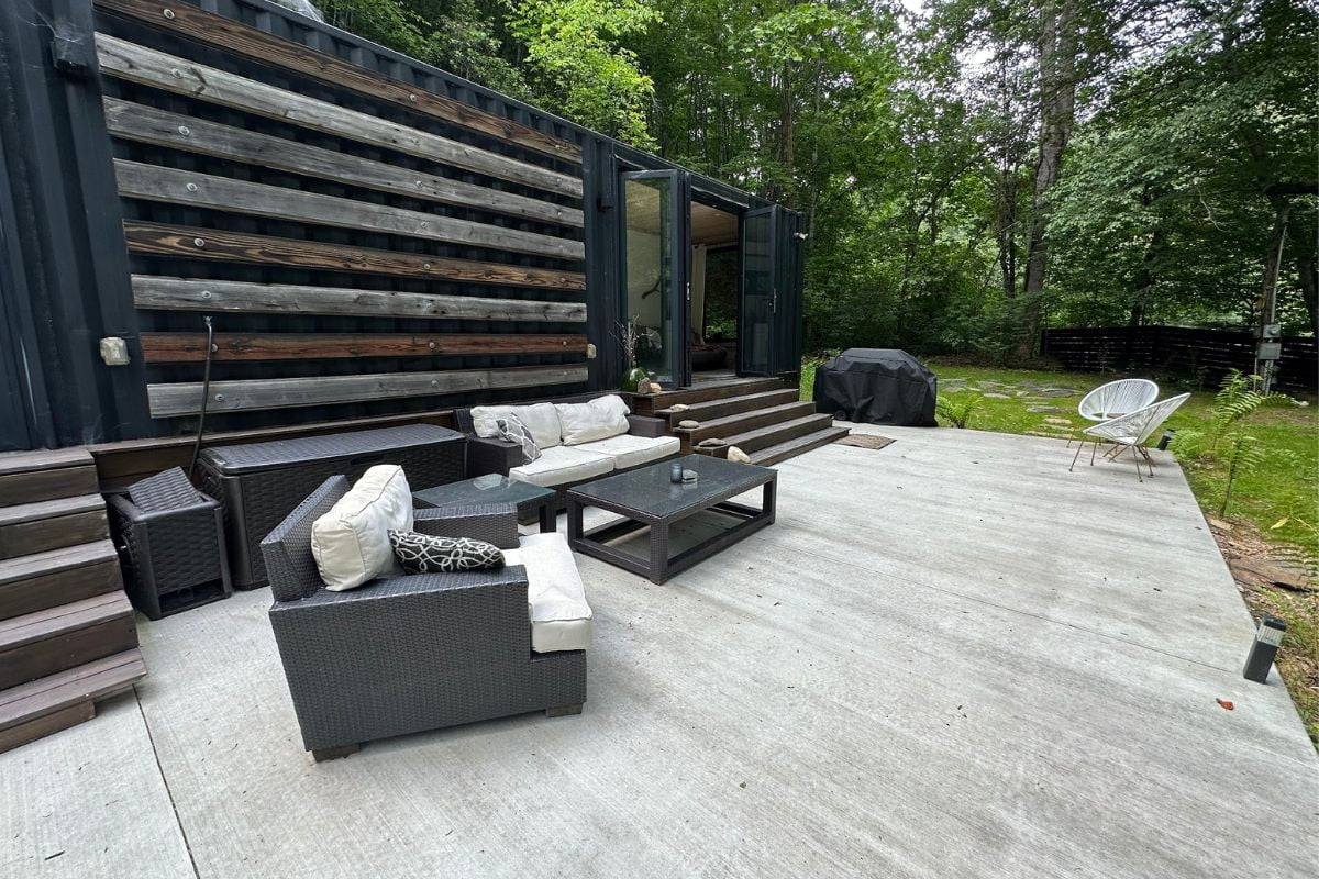 Outdoor patio area with cushioned seating, wicker furniture, a coffee table, and a barbecue grill adjacent to a converted shipping container nestled in a forested area. Perfect for those seeking eco friendly travel tips and sustainable living.