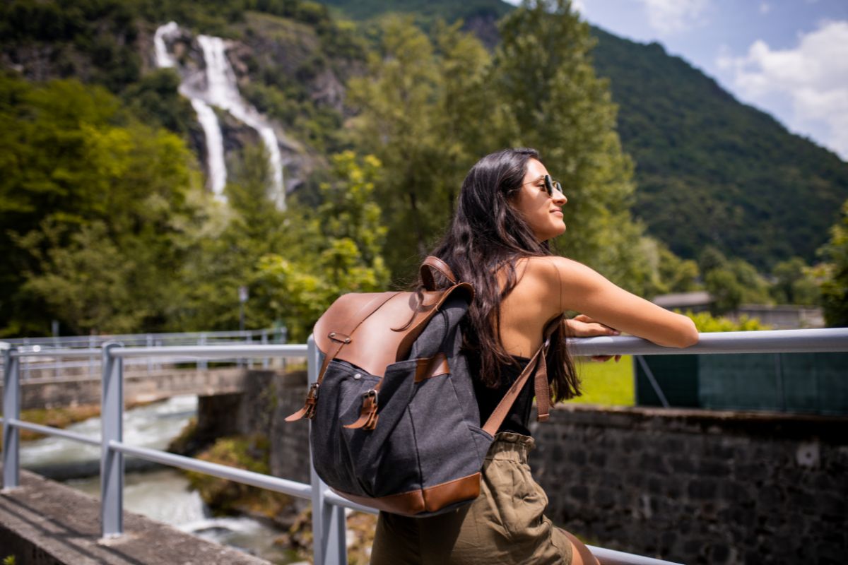 A woman with long hair and sunglasses, carrying a large backpack, leans on a railing in front of a scenic mountain landscape with a waterfall and lush greenery, embracing eco friendly travel tips for her adventures.