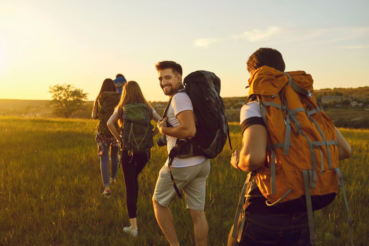 Four people with backpacks walk through a field at sunset, embracing eco friendly travel tips along the way.