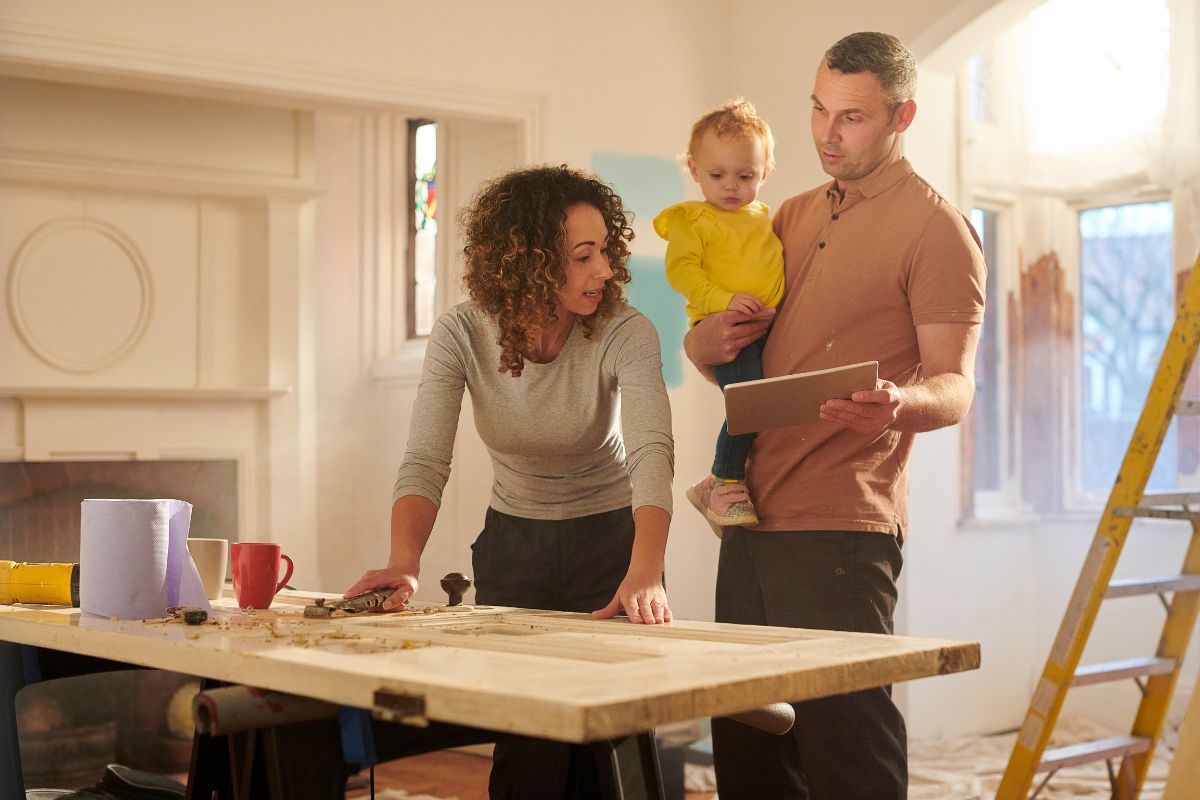 A couple with a child are discussing renovation plans in a partially renovated room. The woman, pointing at a table, shares ideas while the man, holding both a tablet and their child, listens attentively. A ladder in the background hints at ongoing work focused on sustainable living practices.