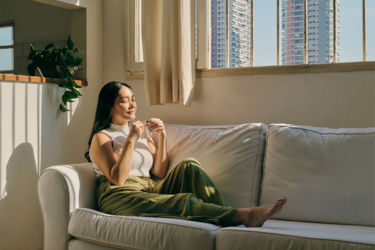 A woman sits on a white couch, eyes closed, holding a mug, with sunlight streaming through the window and tall buildings visible outside—a serene moment embracing sustainable living practices.