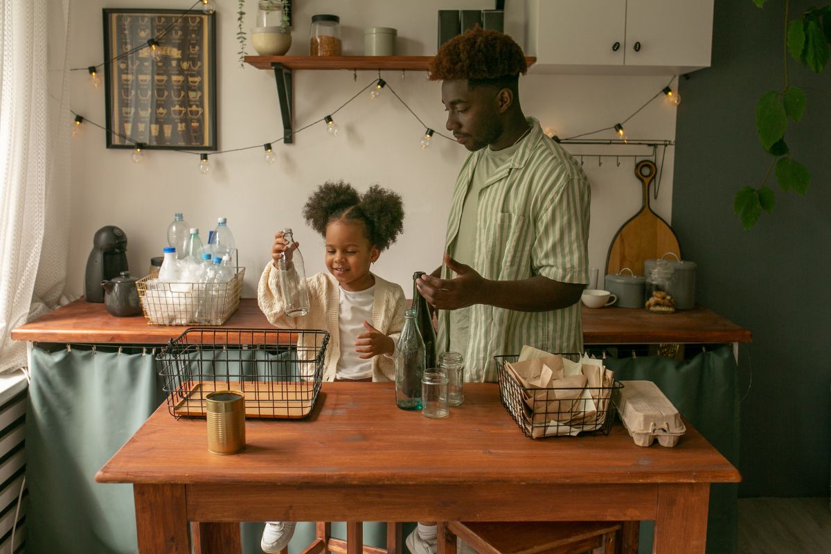 A man and a young girl are in a kitchen arranging items into glass jars and baskets on a wooden table, embracing sustainable living practices. Various jars and bottles are on shelves behind them, with string lights hanging above.